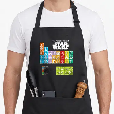 Tablier de cuisine noir avec le tableau périodique des personnages et vaisseaux de Star Wars colorés par faction.