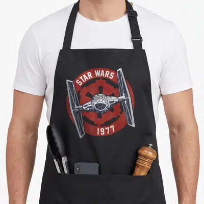 Tablier de cuisine noir avec un TIE Fighter blanc et gris sur un badge circulaire rouge portant les mots Star Wars et 1977.