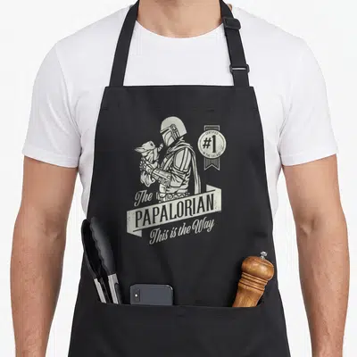 Tablier de cuisine noir avec illustration monochrome du Mandalorien tenant Grogu et le texte The Papalorian This is the Way.