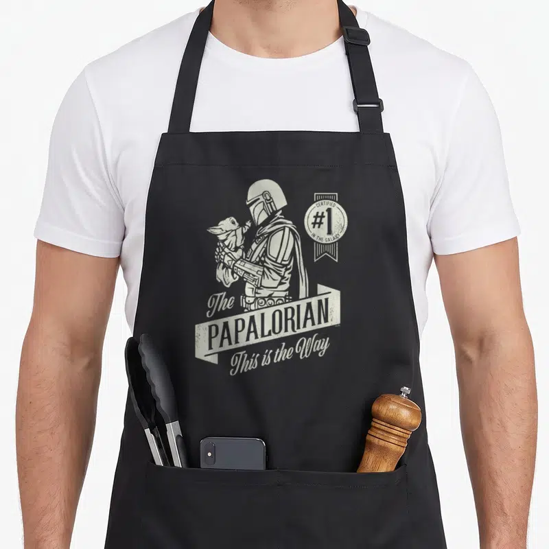 Tablier de cuisine noir avec illustration monochrome du Mandalorien tenant Grogu et le texte The Papalorian This is the Way.