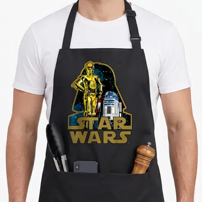 Tablier de cuisine noir avec l'illustration de C-3PO et R2-D2 sous le profil de Dark Vador et le logo Star Wars doré.