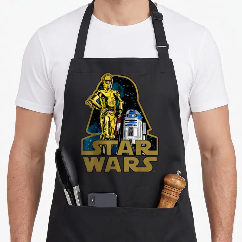 Tablier de cuisine noir avec l'illustration de C-3PO et R2-D2 sous le profil de Dark Vador et le logo Star Wars doré.