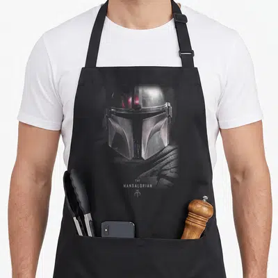 Tablier de cuisine noir avec illustration détaillée du casque argenté du Mandalorian et son logo distinctif en bas.