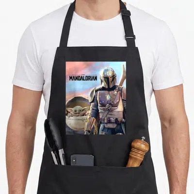 Tablier de cuisine noir avec le Mandalorian et Grogu dans leur brouette sous un ciel rose et bleu.