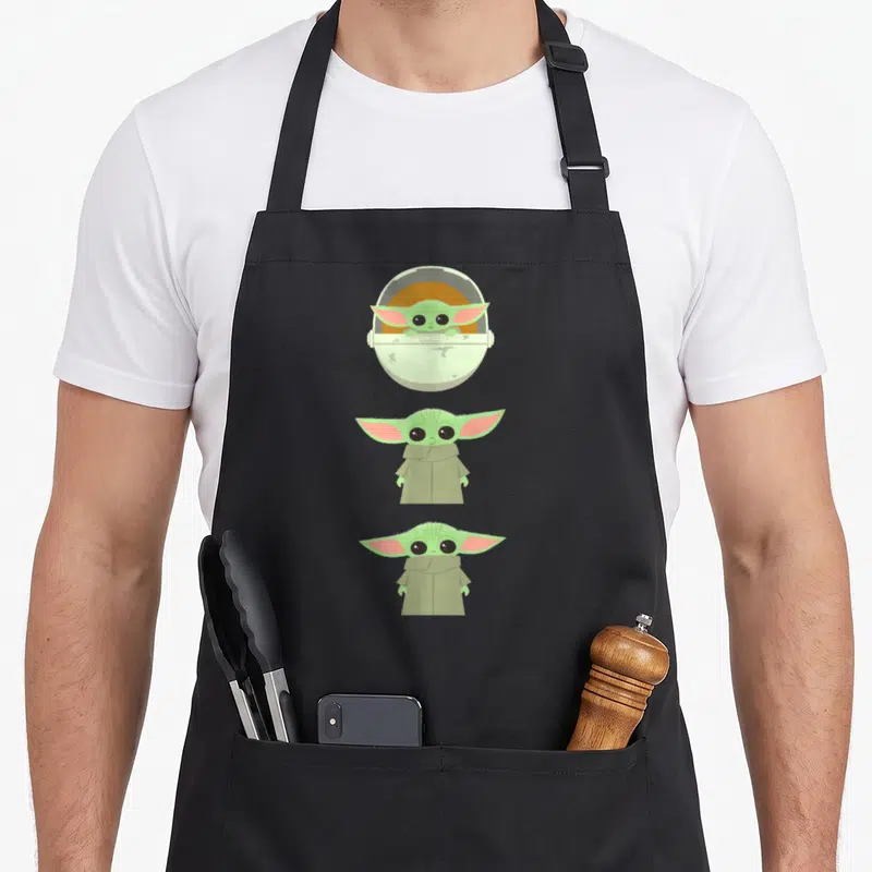 Tablier de cuisine noir avec trois illustrations verticales de Grogu en style cartoon, incluant une version dans son chariot rond et deux versions debout en manteau beige.