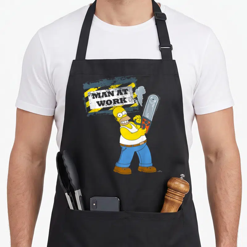 Tablier de cuisine noir avec Homer Simpson tenant une tronçonneuse sous un panneau Man at Work jaune et gris.