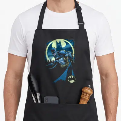 Tablier de cuisine noir avec une illustration de Batman bleu sautant devant un grand logo circulaire jaune et une silhouette de ville en arrière-plan.