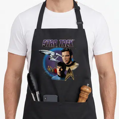 Tablier de cuisine noir avec illustration vintage Star Trek montrant Kirk, Spock et l'Enterprise dans un cercle bleu spatial.