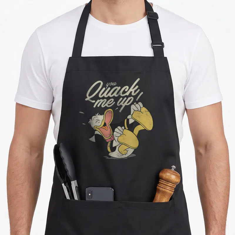 Tablier de cuisine noir avec Donald Duck riant et l'inscription You Quack Me Up en lettres vintage.
