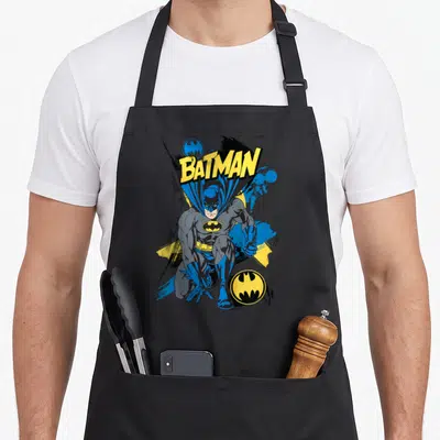 Tablier de cuisine noir avec Batman accroupi, texte jaune et logos circulaires bleus et jaunes.