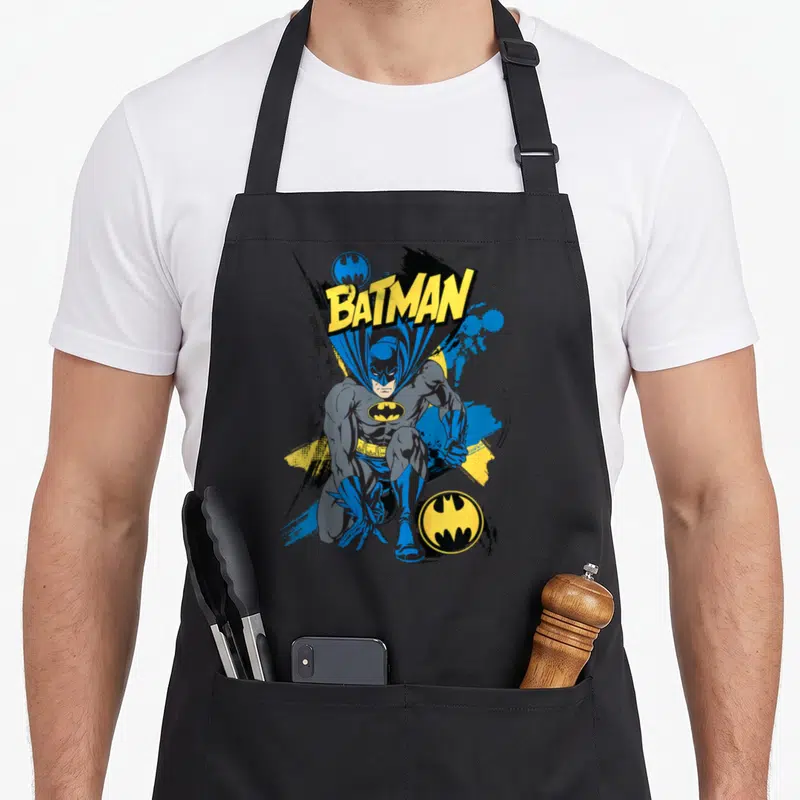 Tablier de cuisine noir avec Batman accroupi, texte jaune et logos circulaires bleus et jaunes.