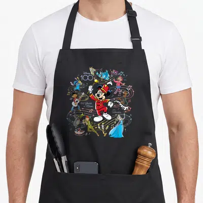 Tablier de cuisine noir avec Mickey en chef d'orchestre entouré de personnages Disney jouant des instruments sur fond de notes de musique.