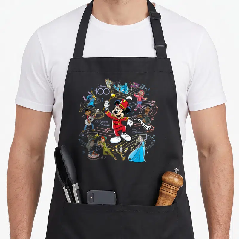 Tablier de cuisine noir avec Mickey en chef d'orchestre entouré de personnages Disney jouant des instruments sur fond de notes de musique.
