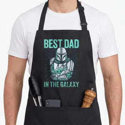 Tablier de cuisine noir avec illustration du Mandalorian tenant Grogu et le texte Best Dad In The Galaxy en turquoise.