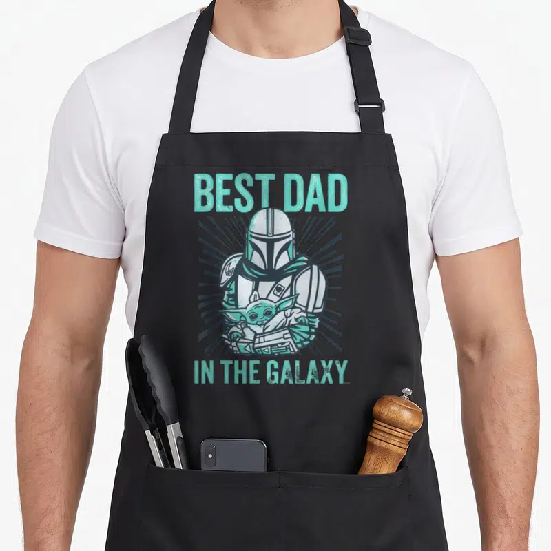Tablier de cuisine noir avec illustration du Mandalorian tenant Grogu et le texte Best Dad In The Galaxy en turquoise.