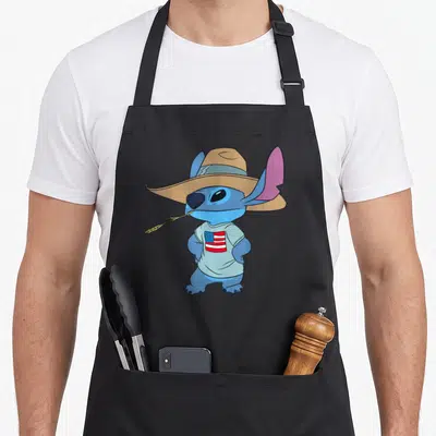 Tablier de cuisine noir avec Stitch bleu coiffé d'un chapeau de cowboy tenant une tige d'herbe dans sa bouche.