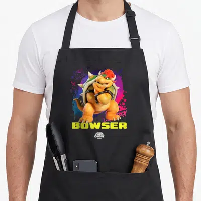 Tablier de cuisine noir avec Bowser le Super Mario Galaxy Movie aux bras croisés sur fond coloré.