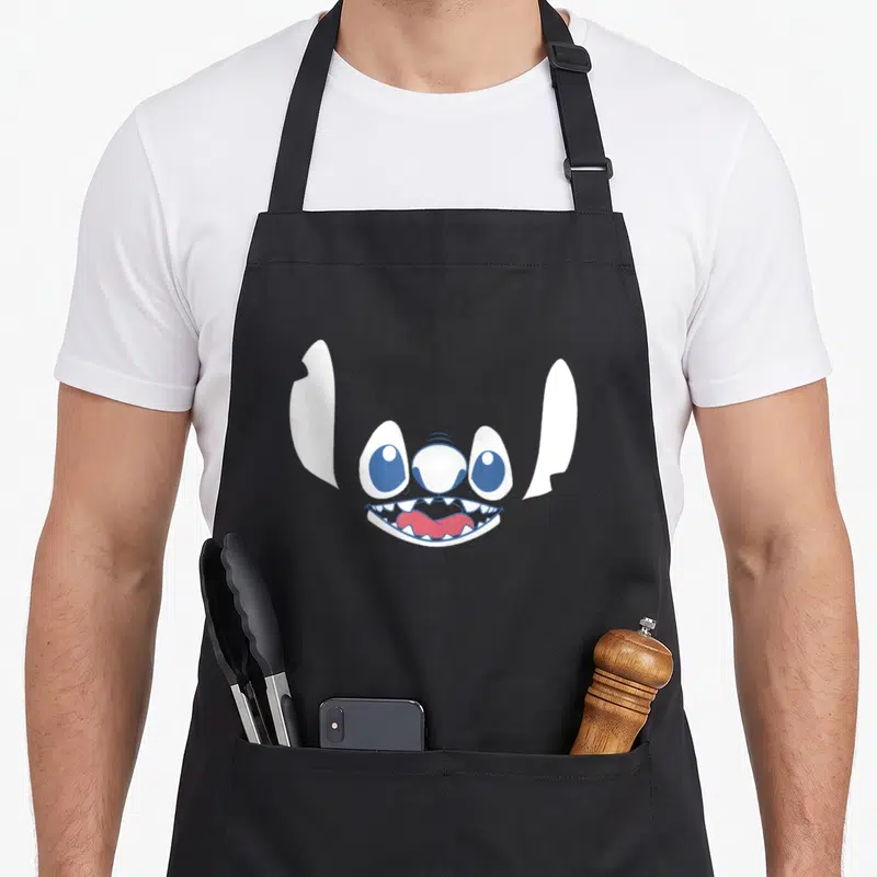 Tablier de cuisine noir avec le visage stylisé de Stitch aux grands yeux bleus et sourire édenté.