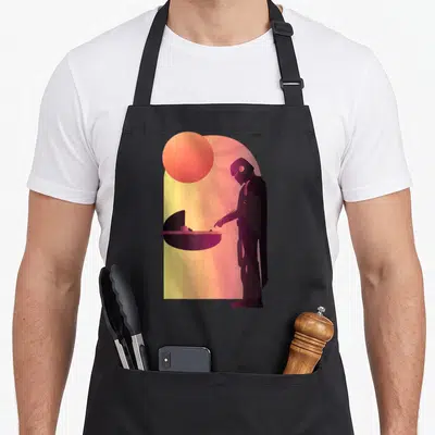 Tablier de cuisine noir avec illustration stylisée du Mandalorien tendant la main vers un berceau sous un soleil orange.