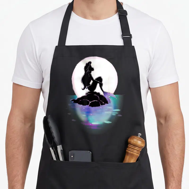 Tablier de cuisine noir avec silhouette sirène assise sur rocher devant lune rose et eau multicolore