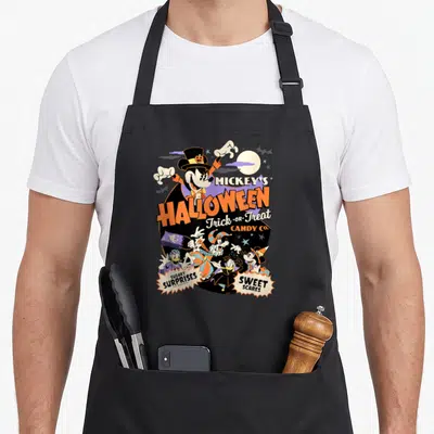 Tablier de cuisine noir avec illustration colorée d'Halloween montrant Mickey et ses amis en tenues festives autour du texte Halloween Trick or Treat Candy Co.