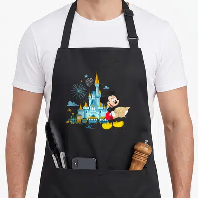 Tablier de cuisine noir avec Mickey Mouse tenant une carte devant un château festif aux couleurs bleues et dorées.