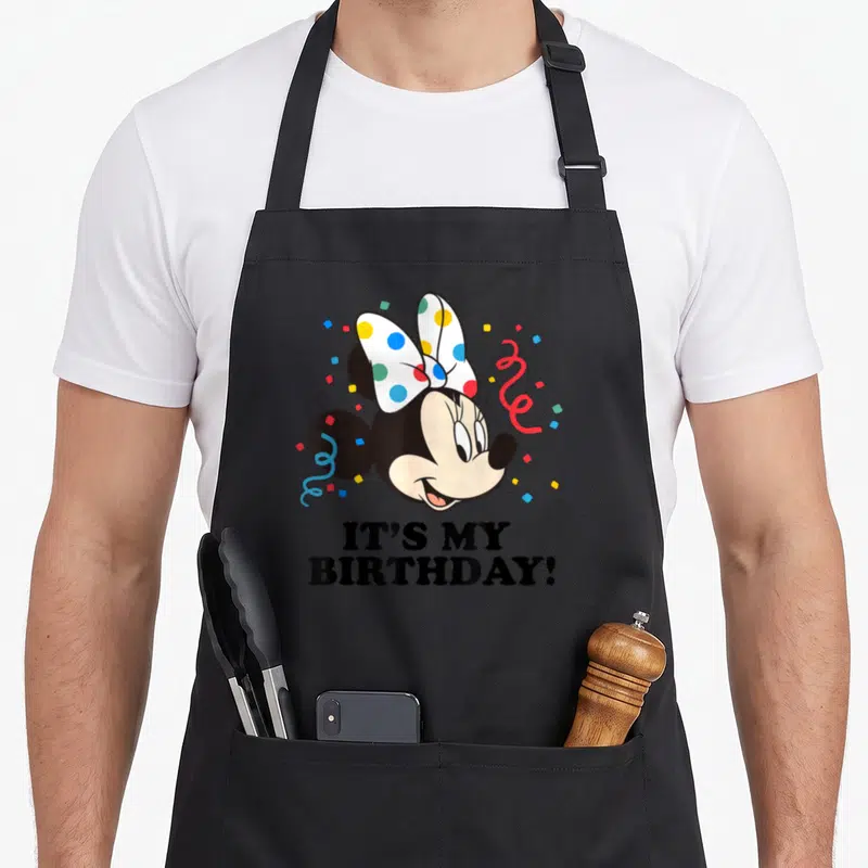 Tablier de cuisine noir avec tête de Minnie Mouse souriante et texte It's My anniversaire entouré de confettis multicolores.