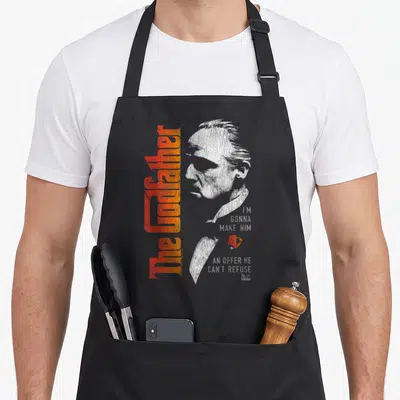 Tablier de cuisine noir avec portrait de Don Vito Corleone et texte The Godfather en orange vertical.
