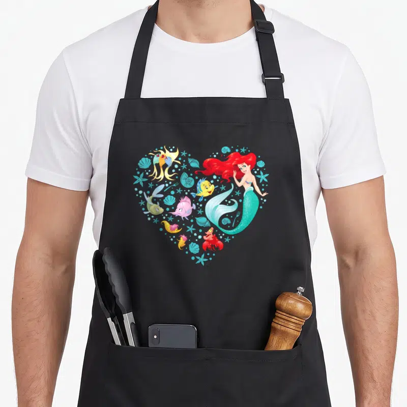 Tablier de cuisine noir avec illustration colorée d'Ariel, Flounder et Sebastian formant un cœur marin.