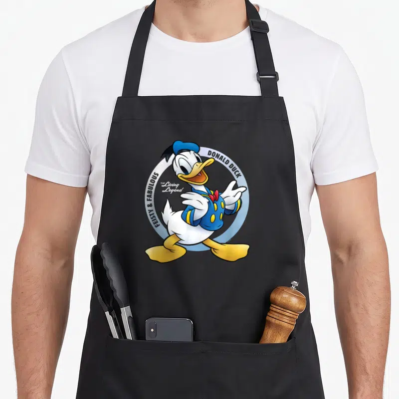 Tablier de cuisine noir avec Donald Duck en tenue classique et texte Feisty & Fabulous The Living Legend dans un cercle bleu clair.