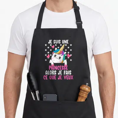 Tablier de cuisine noir avec licorne aux cheveux multicolores, étoiles blanches et coeurs roses entourant le texte imprimé.
