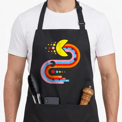 Tablier de cuisine noir avec illustration colorée de Pac-Man dans un labyrinthe de bandes ondulées et de points géométriques.
