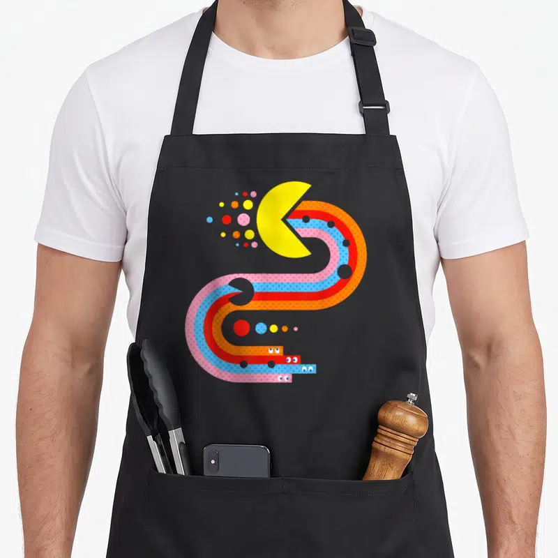 Tablier de cuisine noir avec illustration colorée de Pac-Man dans un labyrinthe de bandes ondulées et de points géométriques.