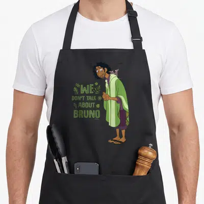Tablier de cuisine noir avec illustration de Bruno en cape verte et texte We Don't Talk About Bruno entouré de motifs végétaux verts.