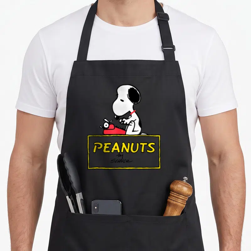 Tablier de cuisine noir avec Snoopy tapant sur une machine rouge et le logo Peanuts by Schulz en dessous.