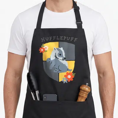 Tablier de cuisine noir avec blaireau stylisé et fleurs sur écu jaune et gris, texte Hufflepuff en haut