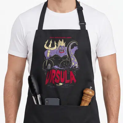 Tablier de cuisine noir avec une illustration colorée d'Ursula tenant un trident doré et son nom en lettres rouges stylisées.
