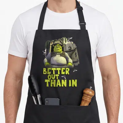 Tablier de cuisine noir avec Shrek debout devant une cabane en bois et le texte Better Out Than In.