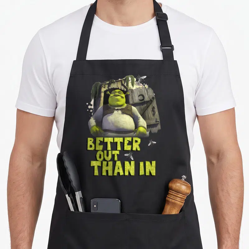 Tablier de cuisine noir avec Shrek debout devant une cabane en bois et le texte Better Out Than In.