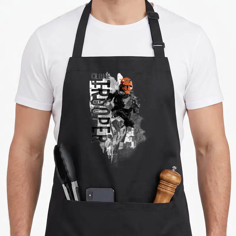 Tablier de cuisine noir avec illustration d'un Clone Trooper casque orange et texte First In Last Standing en style graffiti.