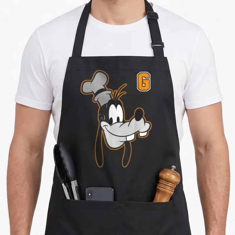 Tablier de cuisine noir avec un grand visage de Goofy portant une toque de chef et une lettre G orange en haut à droite.