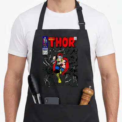 Tablier de cuisine noir avec une illustration colorée de Thor en pleine action sur fond de cases de comics en noir et blanc.
