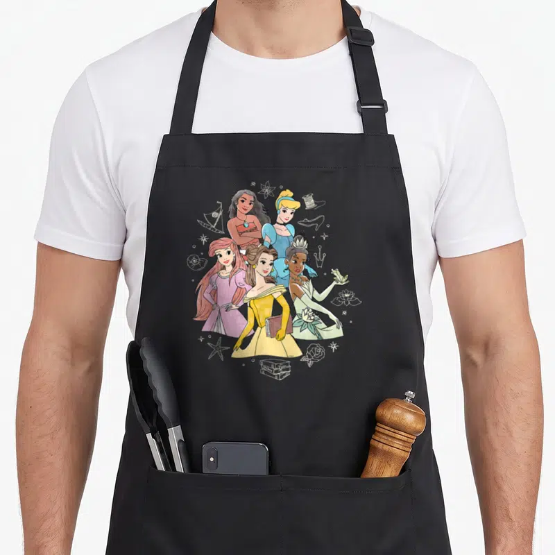 Tablier de cuisine noir avec illustration de cinq princesses en style esquisse au crayon, entourées d'icônes symboliques comme une couronne, un livre et une grenouille.