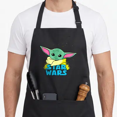 Tablier de cuisine noir avec illustration colorée de Grogu et logo Star Wars en lettres bleues sur fond jaune éclatant.