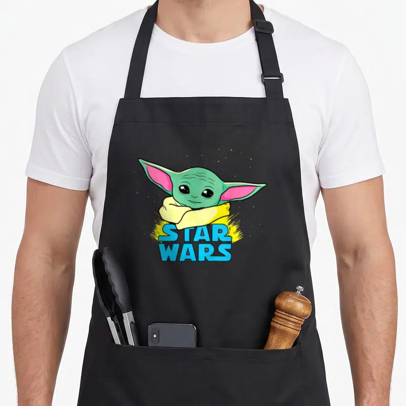 Tablier de cuisine noir avec illustration colorée de Grogu et logo Star Wars en lettres bleues sur fond jaune éclatant.