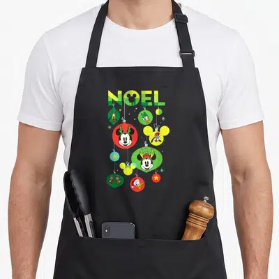 Tablier de cuisine noir avec des boules de Noël colorées portant les visages de Mickey, Donald et Minnie sous le mot NOËL.