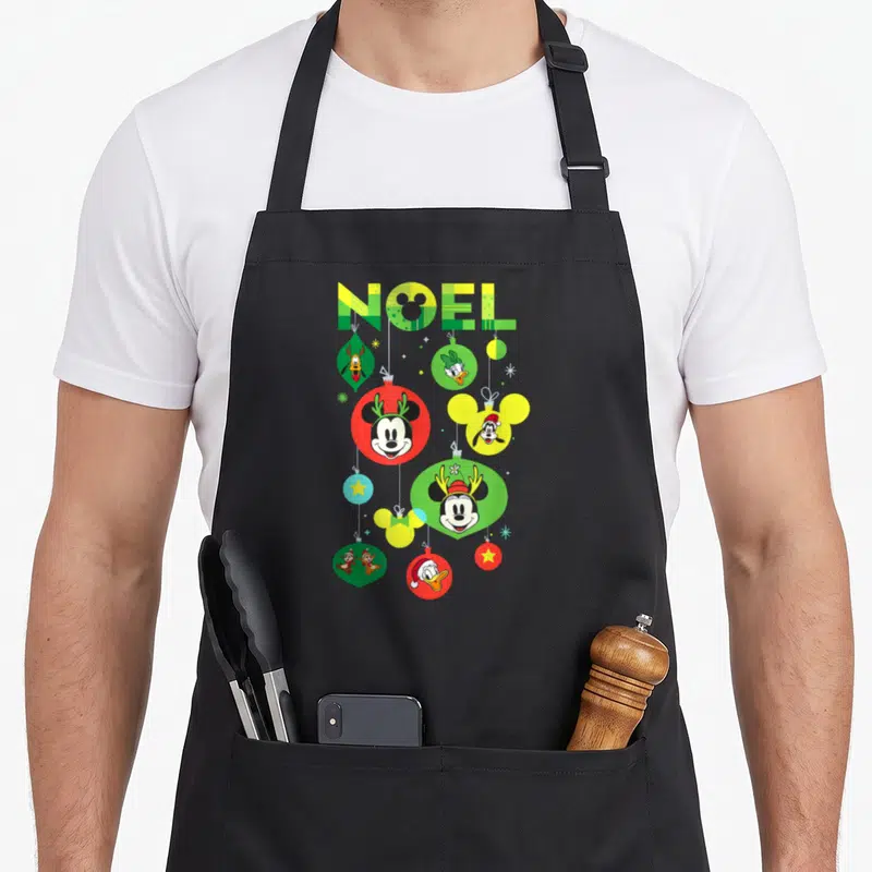 Tablier de cuisine noir avec des boules de Noël colorées portant les visages de Mickey, Donald et Minnie sous le mot NOËL.