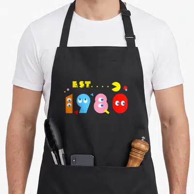 Tablier de cuisine noir avec Est 1980 et personnages de jeu vidéo colorés sur fond uni.