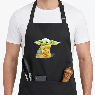 Tablier de cuisine noir avec illustration aquarellée de Grogu tenant une tasse de soupe verte.