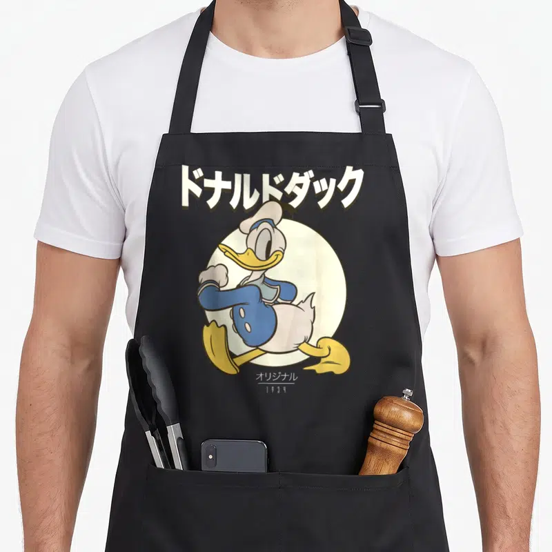 Tablier de cuisine noir avec illustration de Donald Duck en pose dynamique sous des caractères japonais blancs sur fond circulaire crème.
